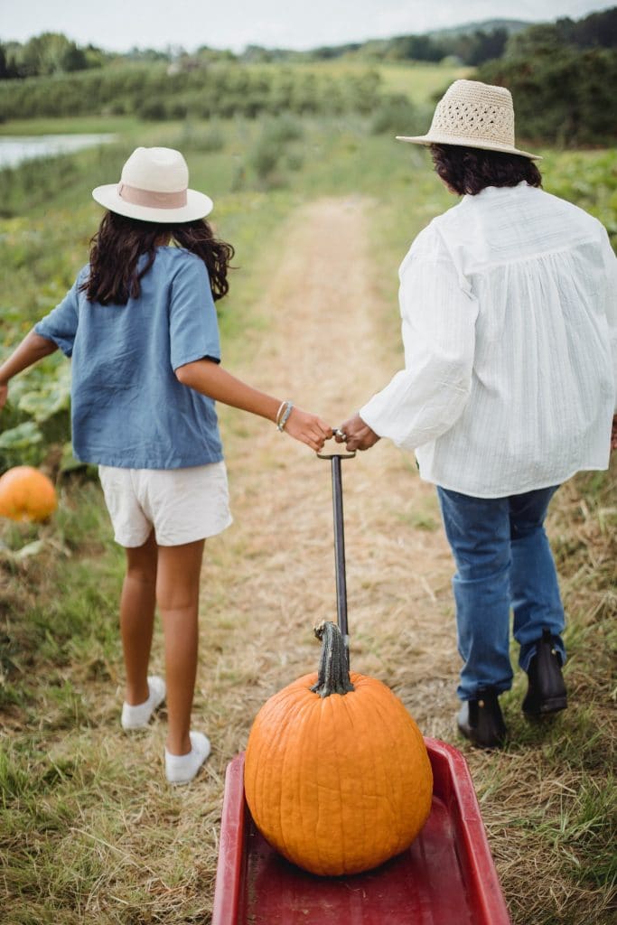 woman and young lady with their backs to camera. Walking away pulling a wagon with orange pumpkins.