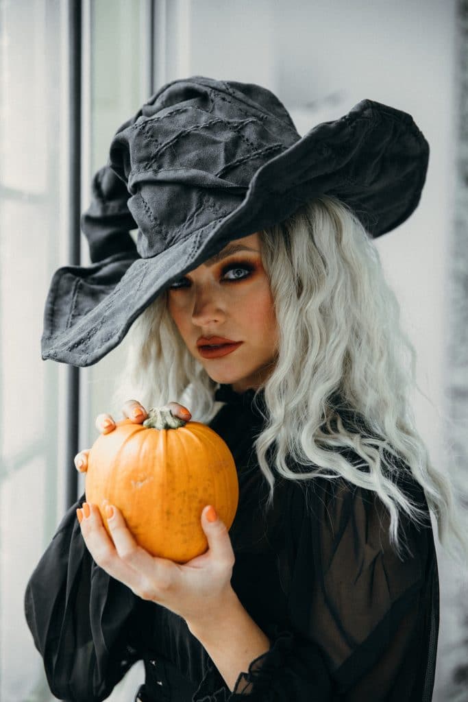 grey haired witch with black hat and black dress holding a small orange pumpkin
