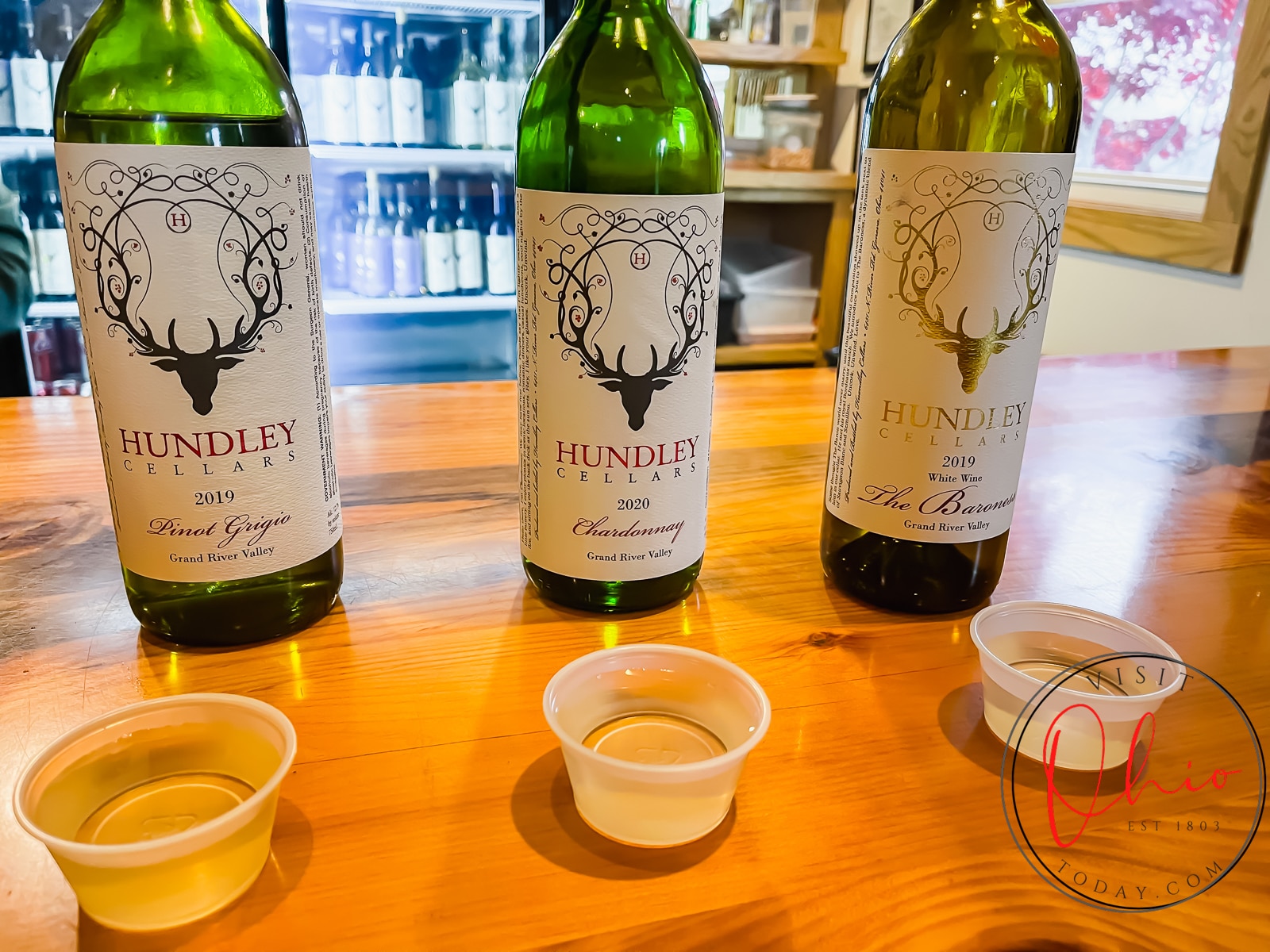 three green wine bottles with off white labels and deer antlers on it, with three clear plastic cups (small) filled with yellow liquid