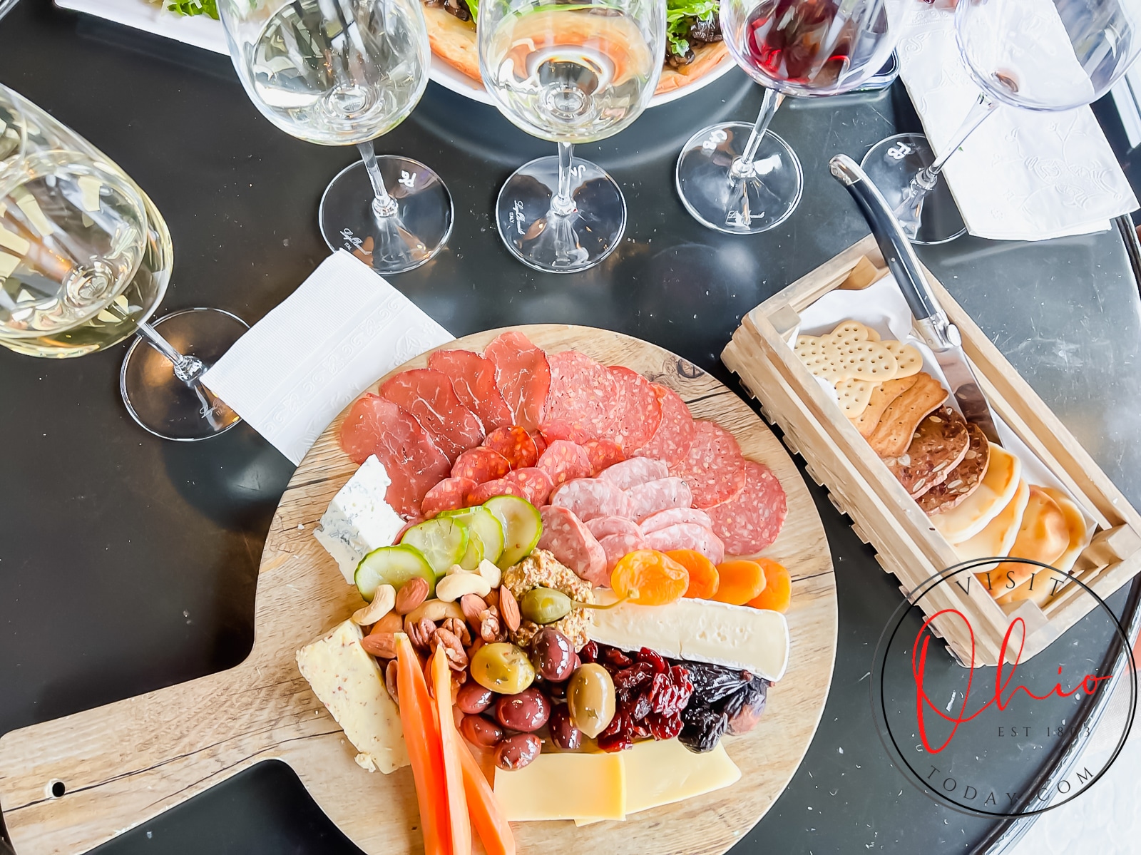 black table, four wine glasses, round wooden board full of veggies, cheese, meats, olives, nuts and a rectangle box of crackers Photo credit: Cindy Gordon of VisitOhioToday.com