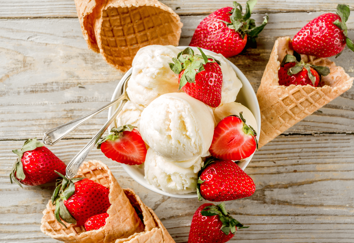 horizontal photo of an overhead shot of a bowl of ice cream scoops with cones around the outside with strawberries on the ice cream, in the cones, and on the wooden surface