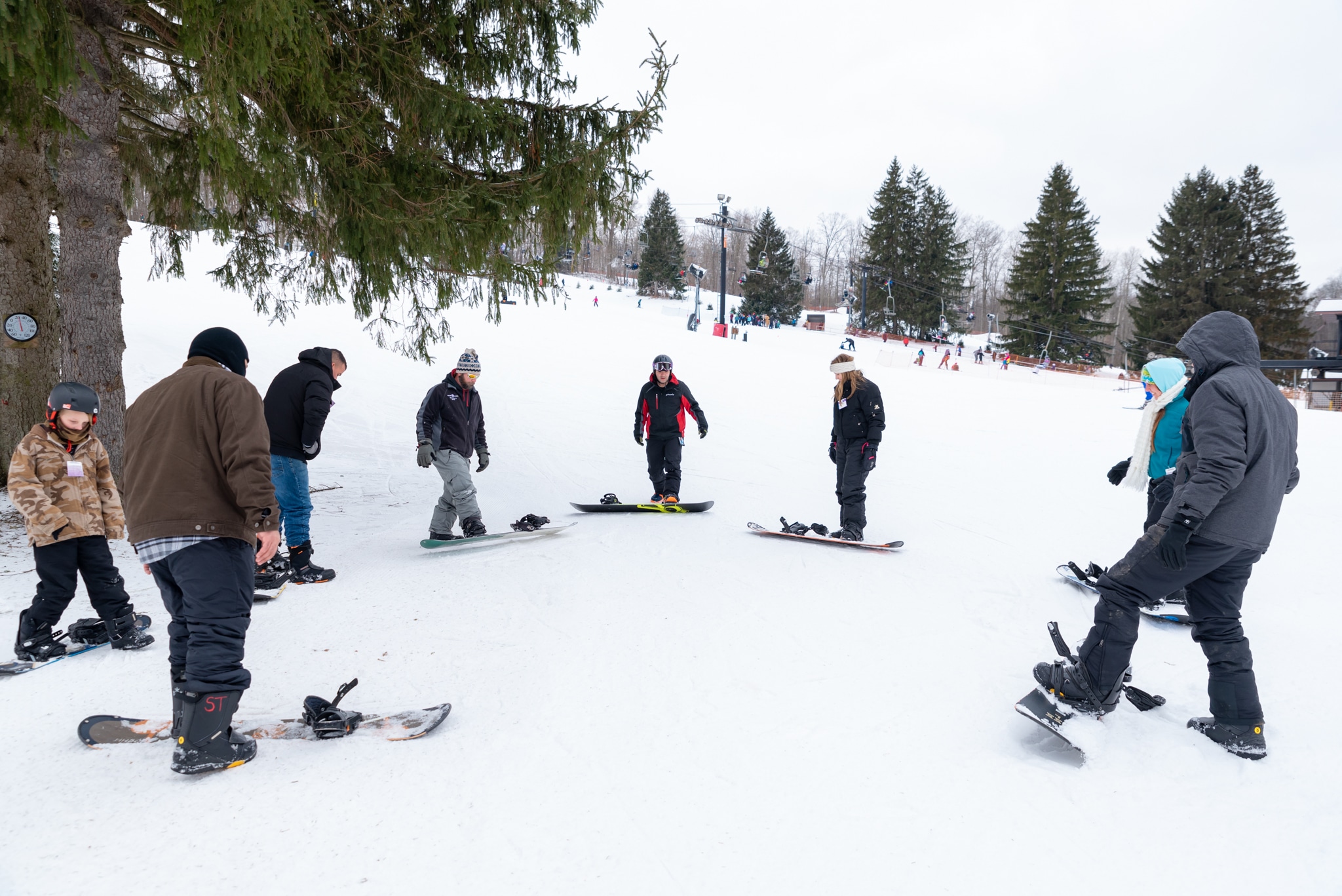 7 people standing on snowboards in a semi circle at snow trails, learning to snow board