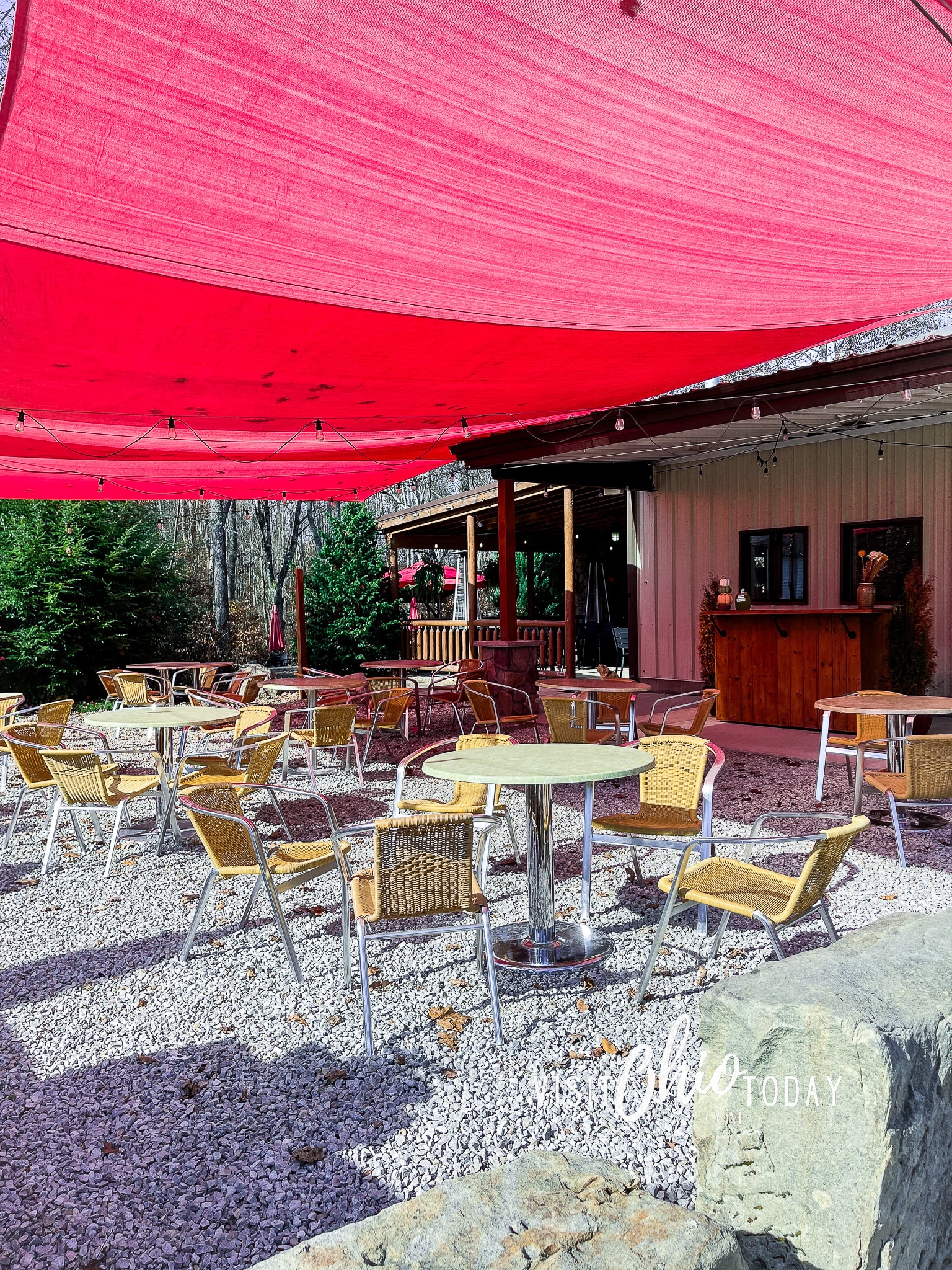 vertical photo of the patio area at Hocking Hills Winery with a red canopy, round tables and wicker chairs. Photo credit: Cindy Gordon of VisitOhioToday.com