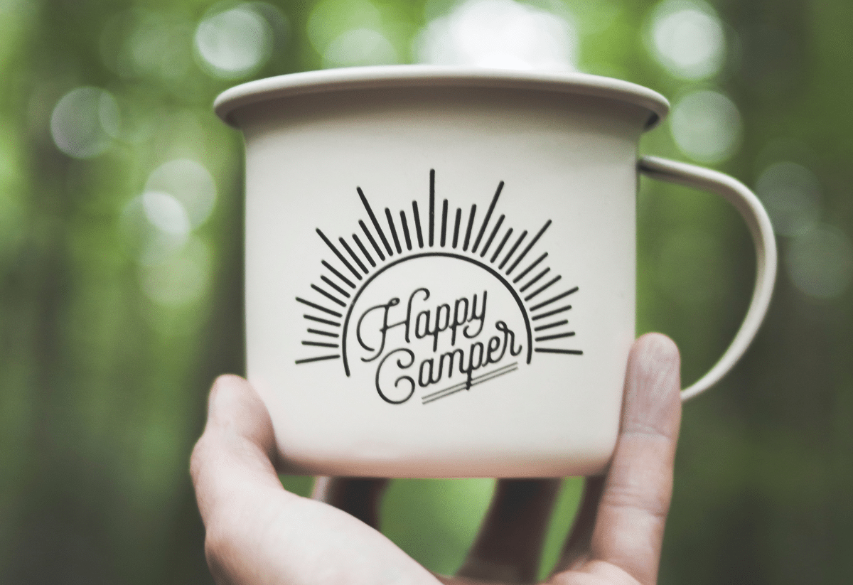 horizontal photo of a hand holding a coffee mug in the air. The mug has the text Happy Camper. The background is blurred foliage