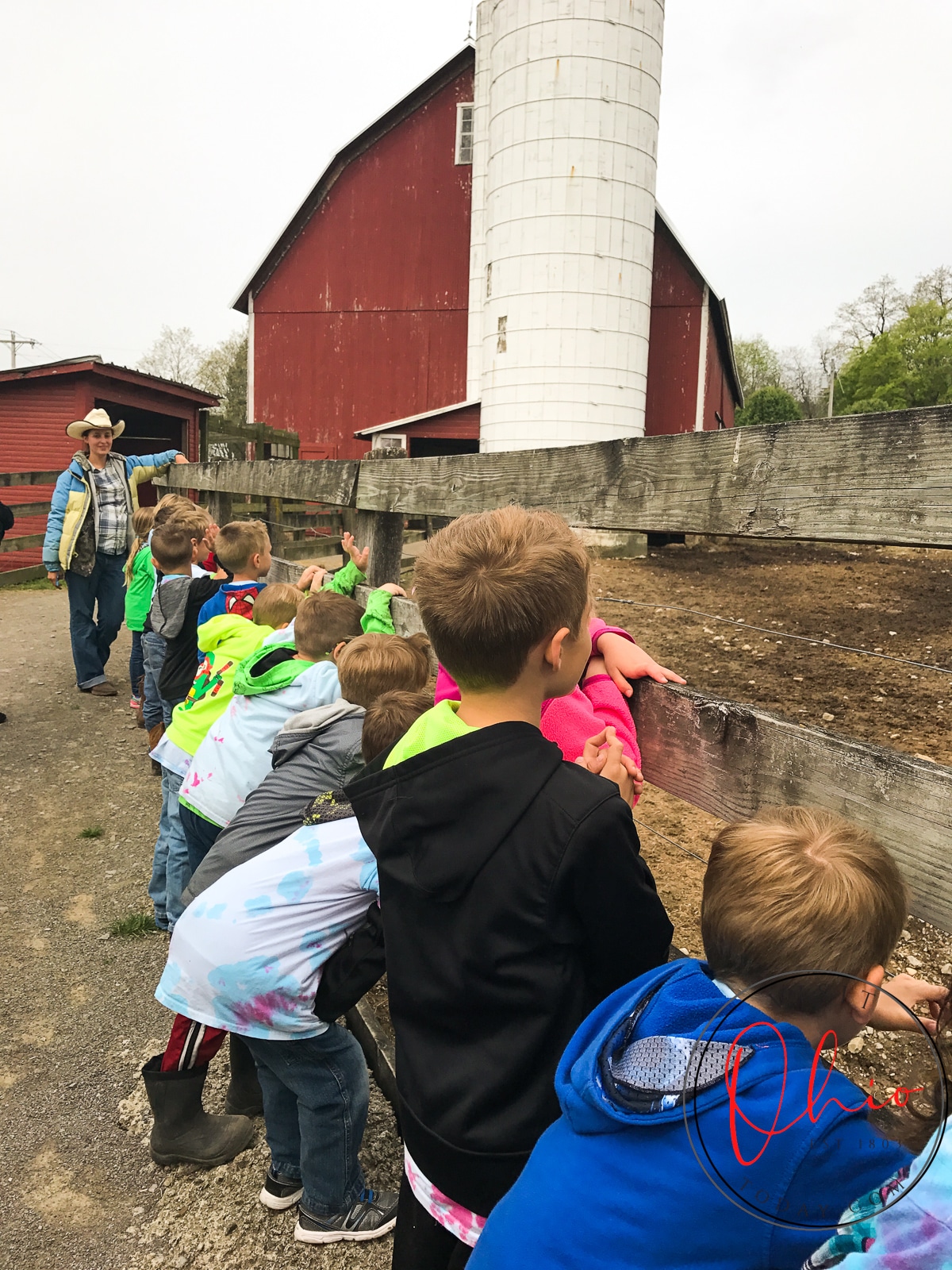 young kids lined up to a wooden fence in front of an old red barn with a white silo