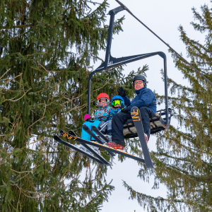 man and two children on ski chair left up high looking down at camera. Evergreens are behind them