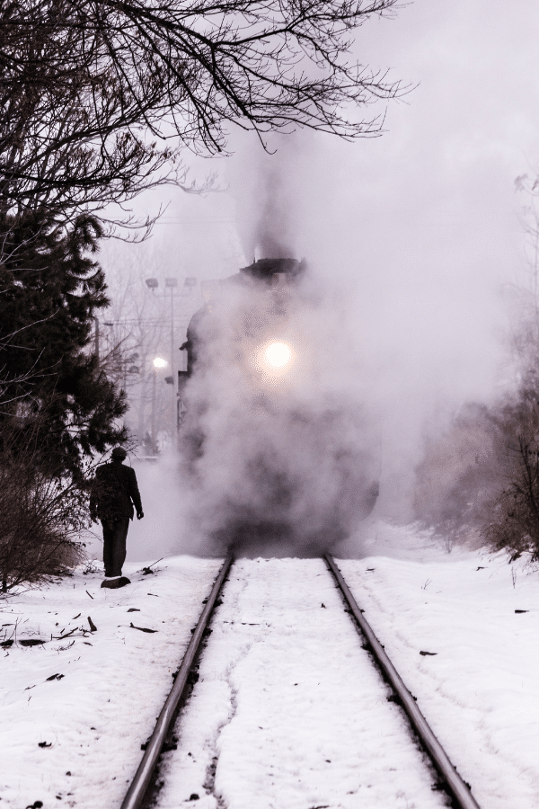 Dark train coming down a snow covered track with fog or steam in the air covering the front of the train. Man dressed in black to the left
