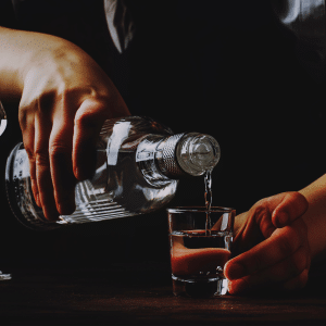 darkly shaded picture. man pouring clear liquid from a glass bottle into a shot glass