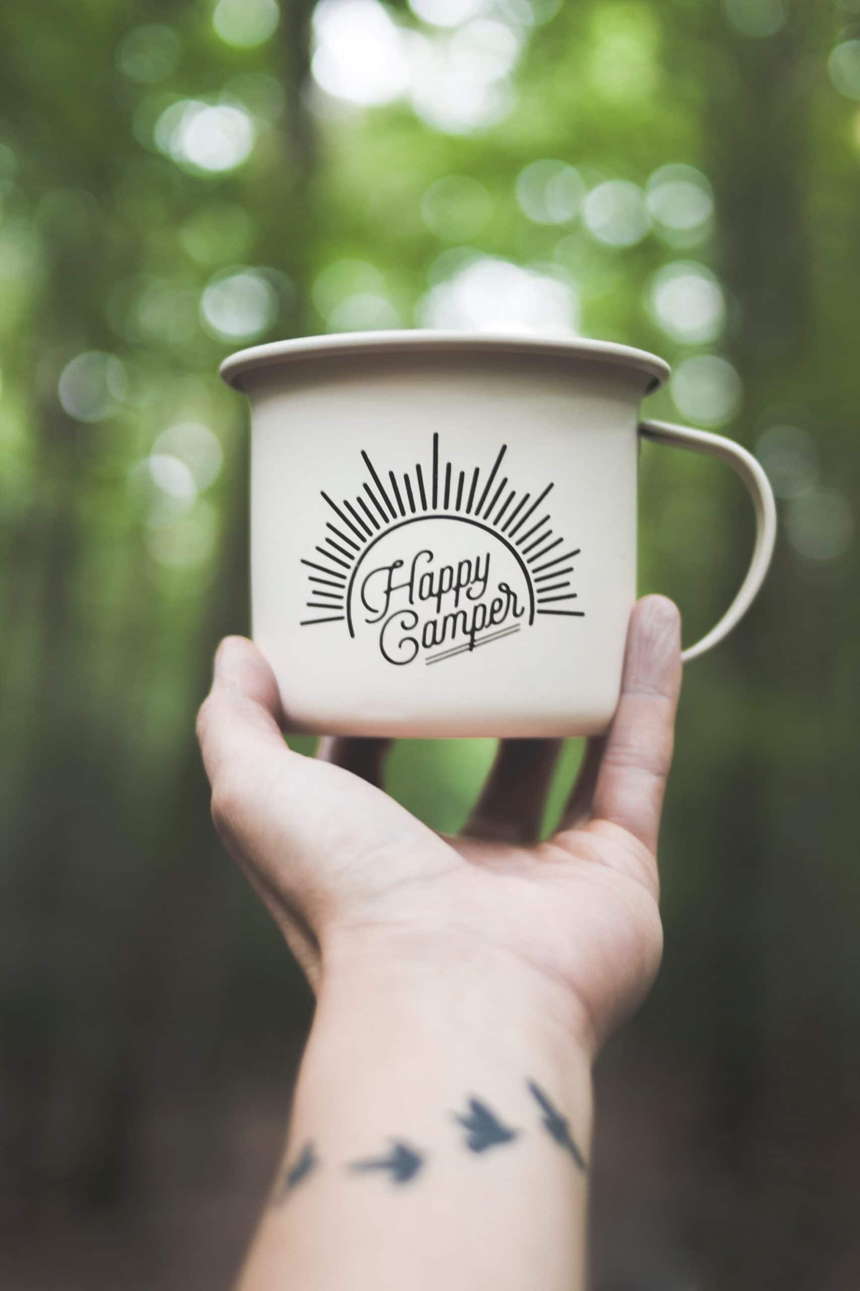 outreached arm holding a tin mug that says happy camper in front of a green forest