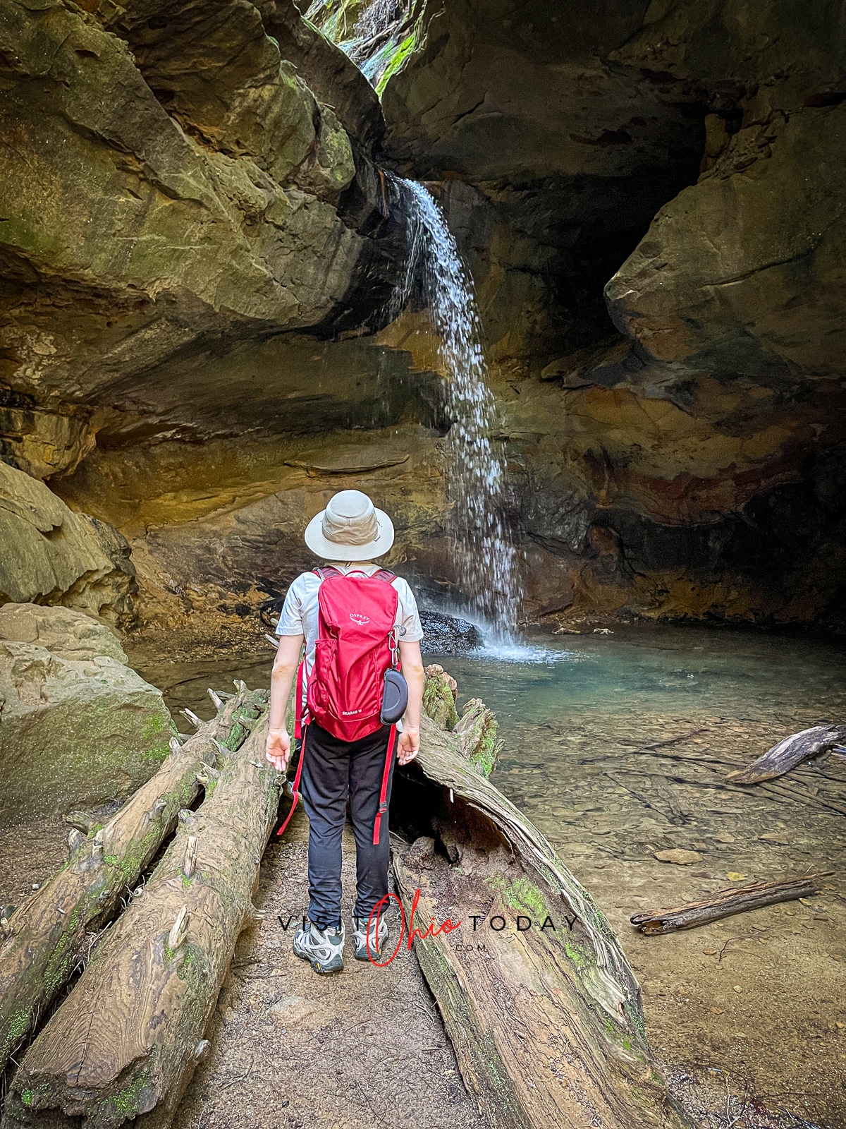 man with hat and red backpack facing a rocking waterfall