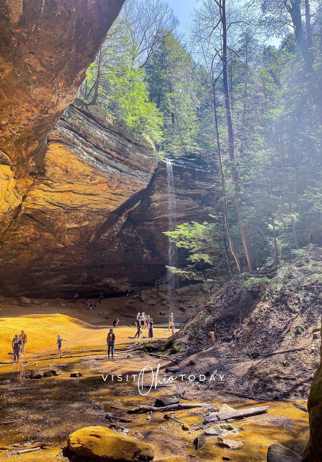 brown cave with tall water fall on right hand side and people walking below the waterfall Photo credit: Cindy Gordon of VisitOhioToday.com