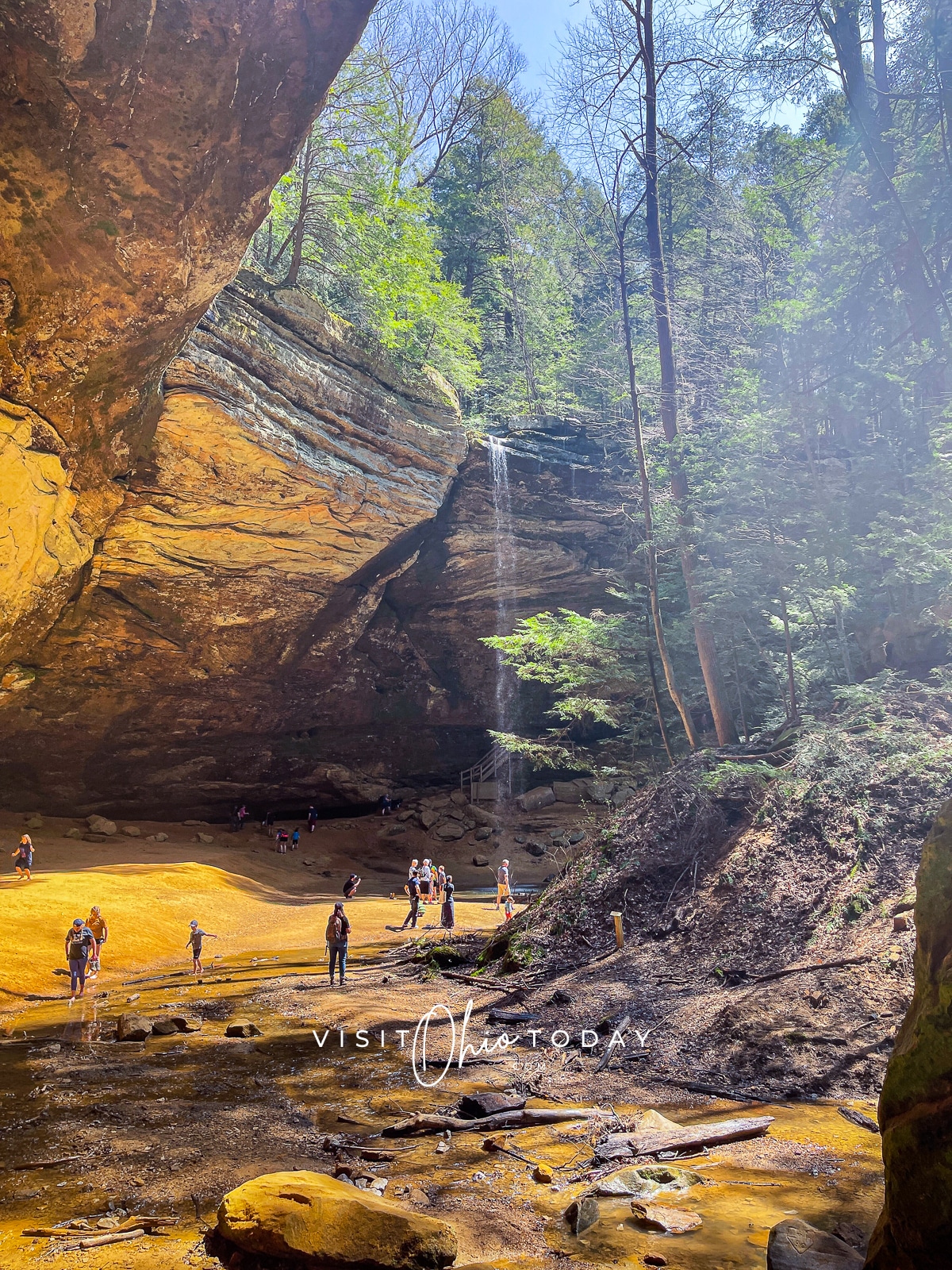 brown cave with tall water fall on right hand side and people walking below the waterfall