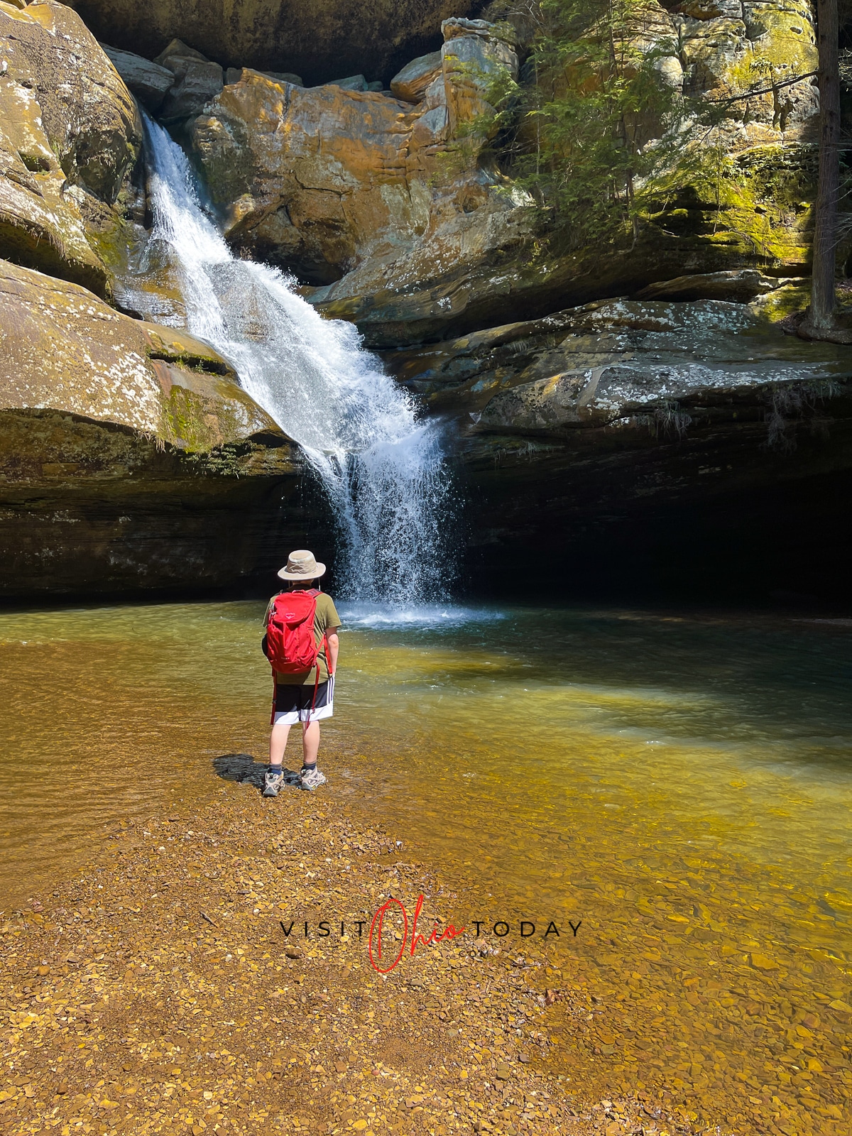 unobstructed view of cedar falls waterfall, water flowing down rocks into a clear pool, person with shorts, a red backpack and hat standing facing the waterfall