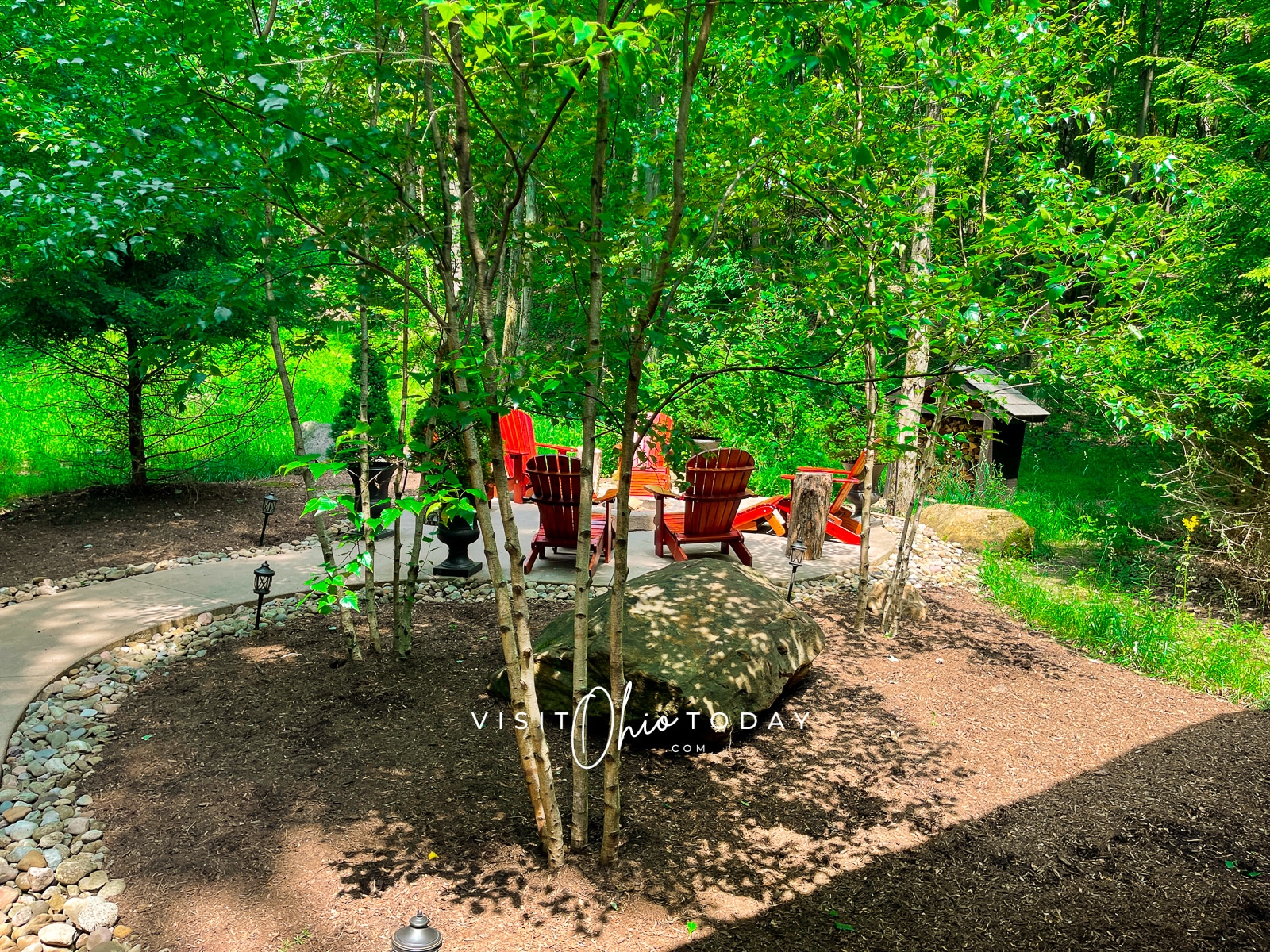 green trees and paved path leading to fire pit with 6 red chairs