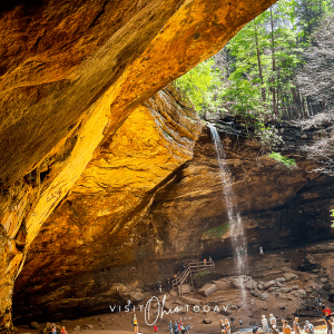 brown cave with tall water fall on right hand side and people walking below the waterfall