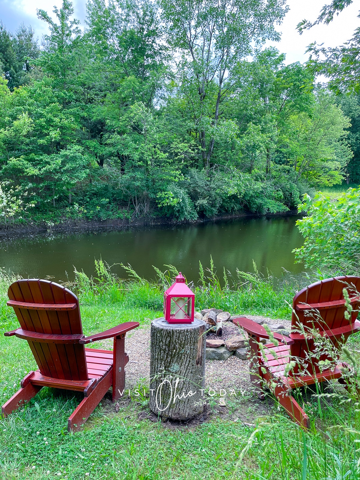 Two red chairs facing a firepit and a lake. There is a red lantern on a log between the chairs. 