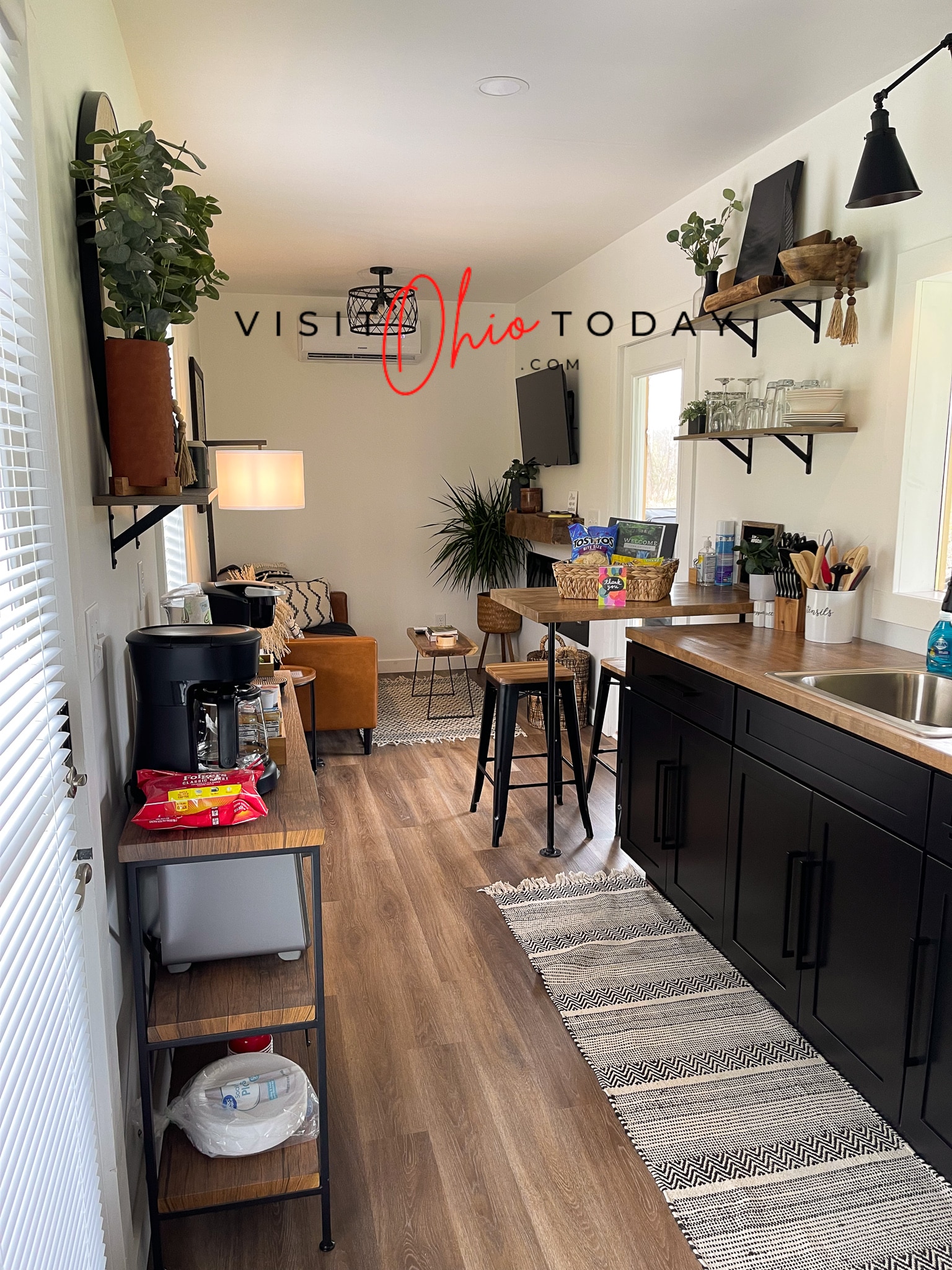 long skinny view of tiny house kitchen and living room. Can see counter, a stool, plant couch, shelf with coffee station Photo credit: Cindy Gordon of VisitOhioToday.com