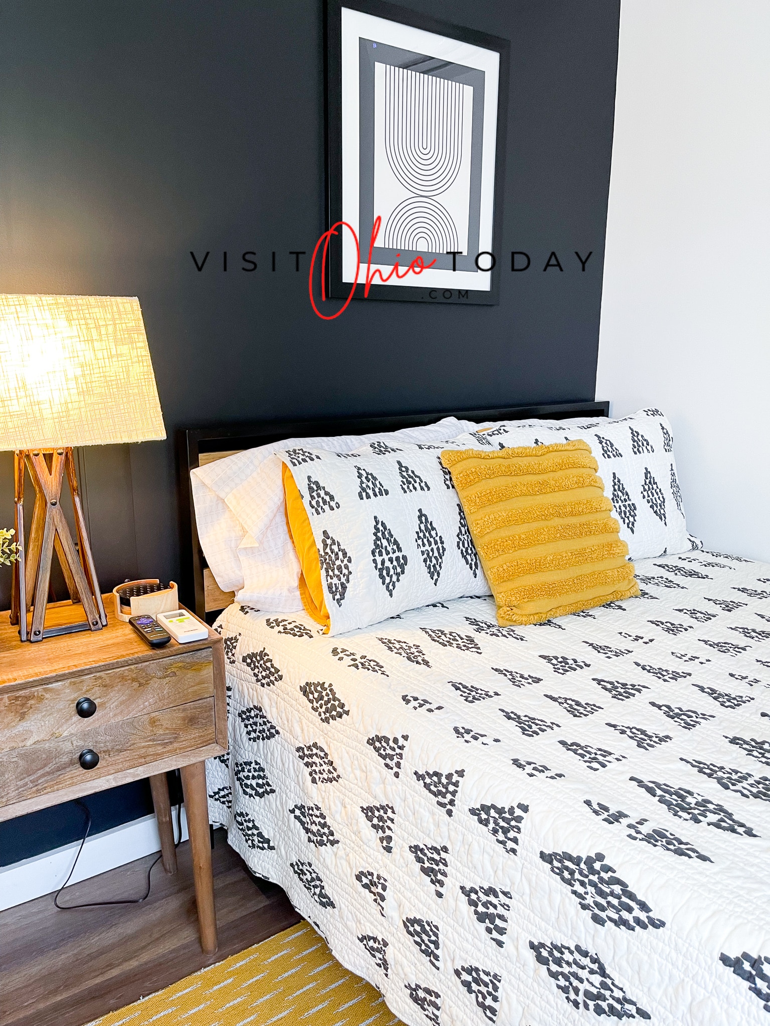 lamp on bedside table, bed with black and white comforter and yellow pillow, black wall with art hung