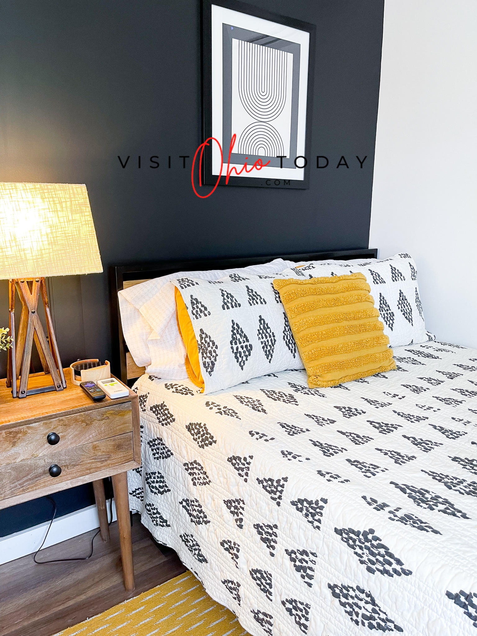 lamp on bedside table, bed with black and white comforter and yellow pillow, black wall with art hung Photo credit: Cindy Gordon of VisitOhioToday.com