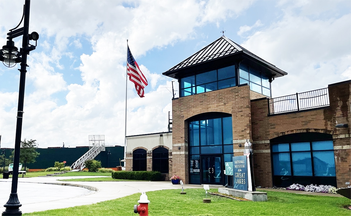 horizontal photo of the National Museum of the Great Lakes with a lamp post and grass in the foreground and a large ship in the background. Image credit: Mbrickn, CC0, via Wikimedia Commons