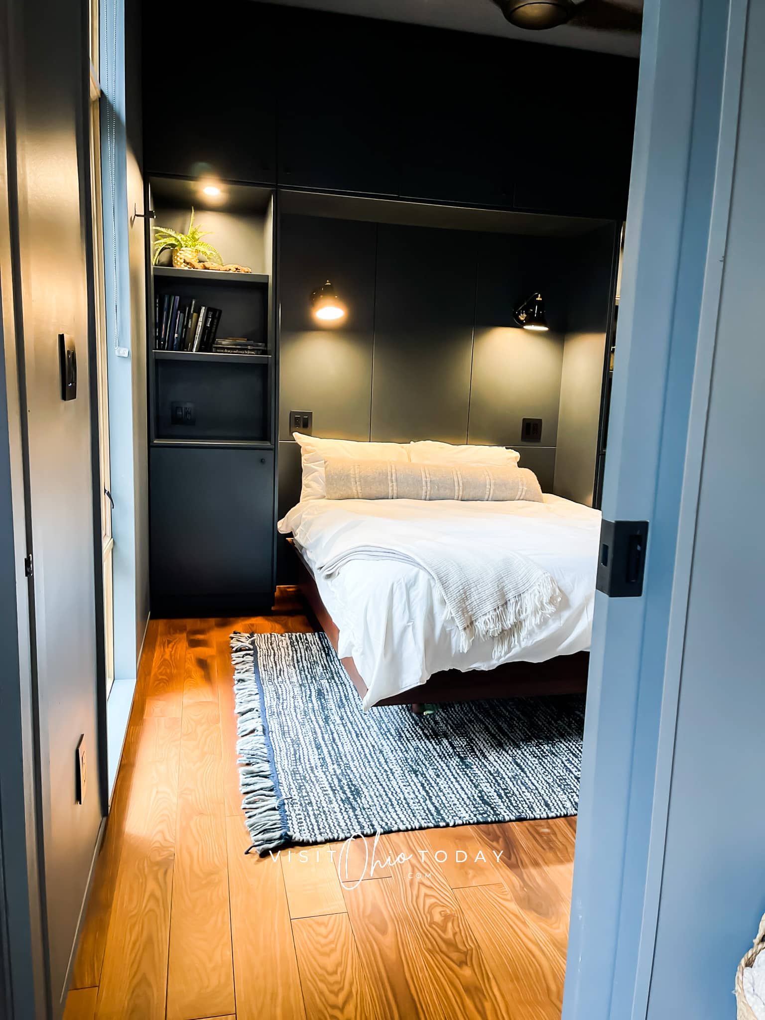 Pictured is a bed with white covers a black and white rug under it and wall lights