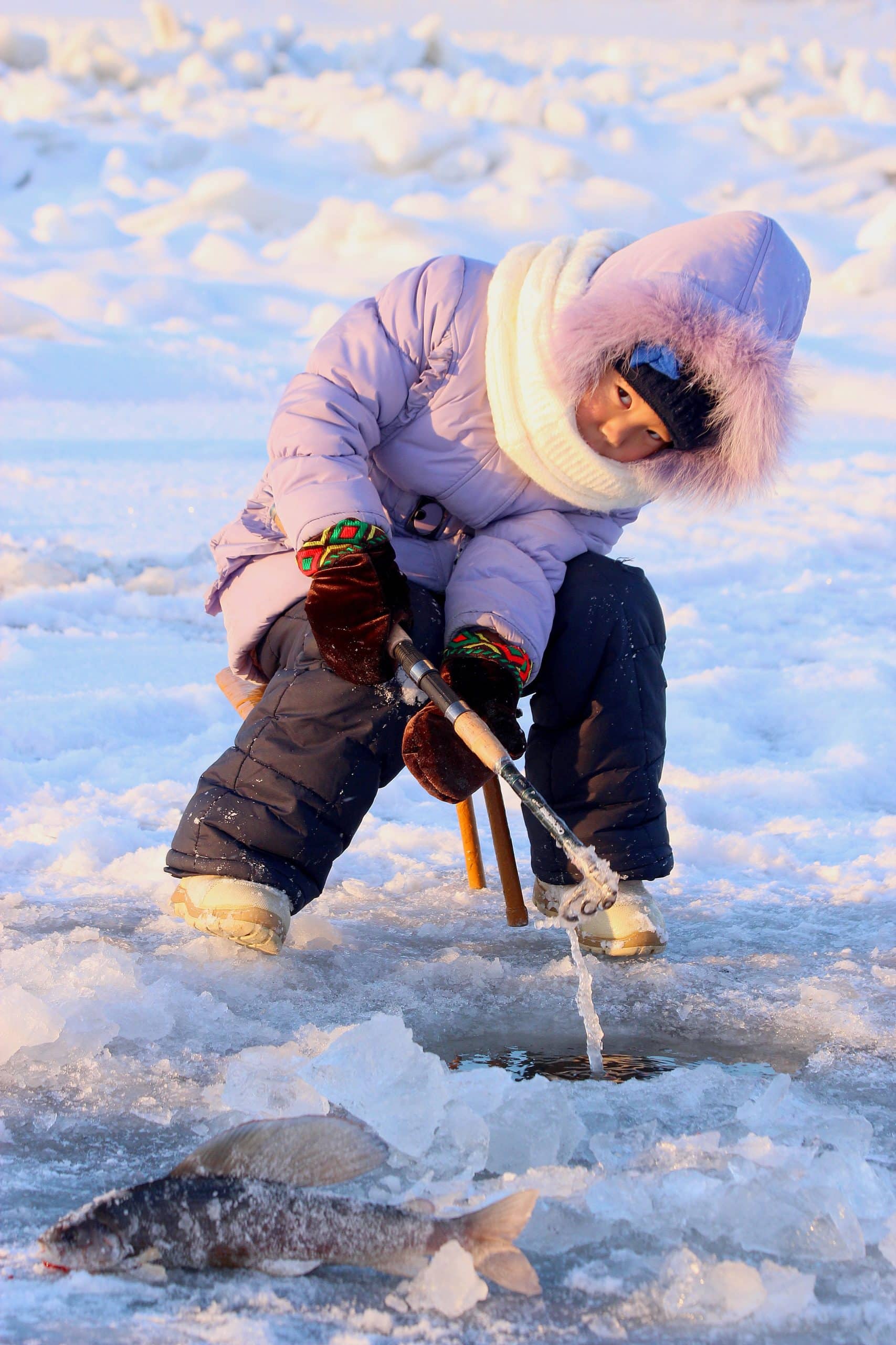 A child dressed in a pink winter coat and cream coloured scarf ice fishing
