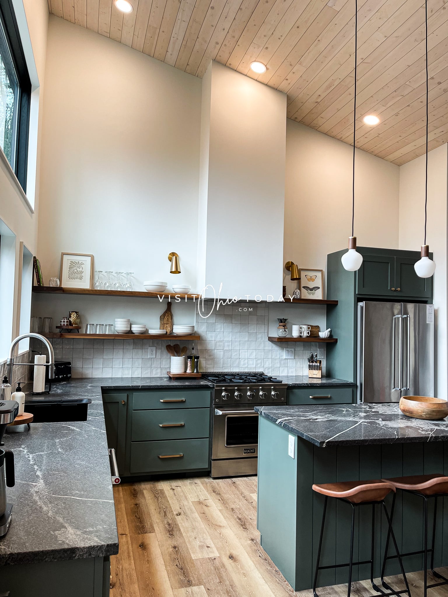 kitchen with white and tileed white wall, green cabinets, black counter tops, silver gas stove in middle and floating brown wooden shelves Photo credit: Cindy Gordon of VisitOhioToday.com