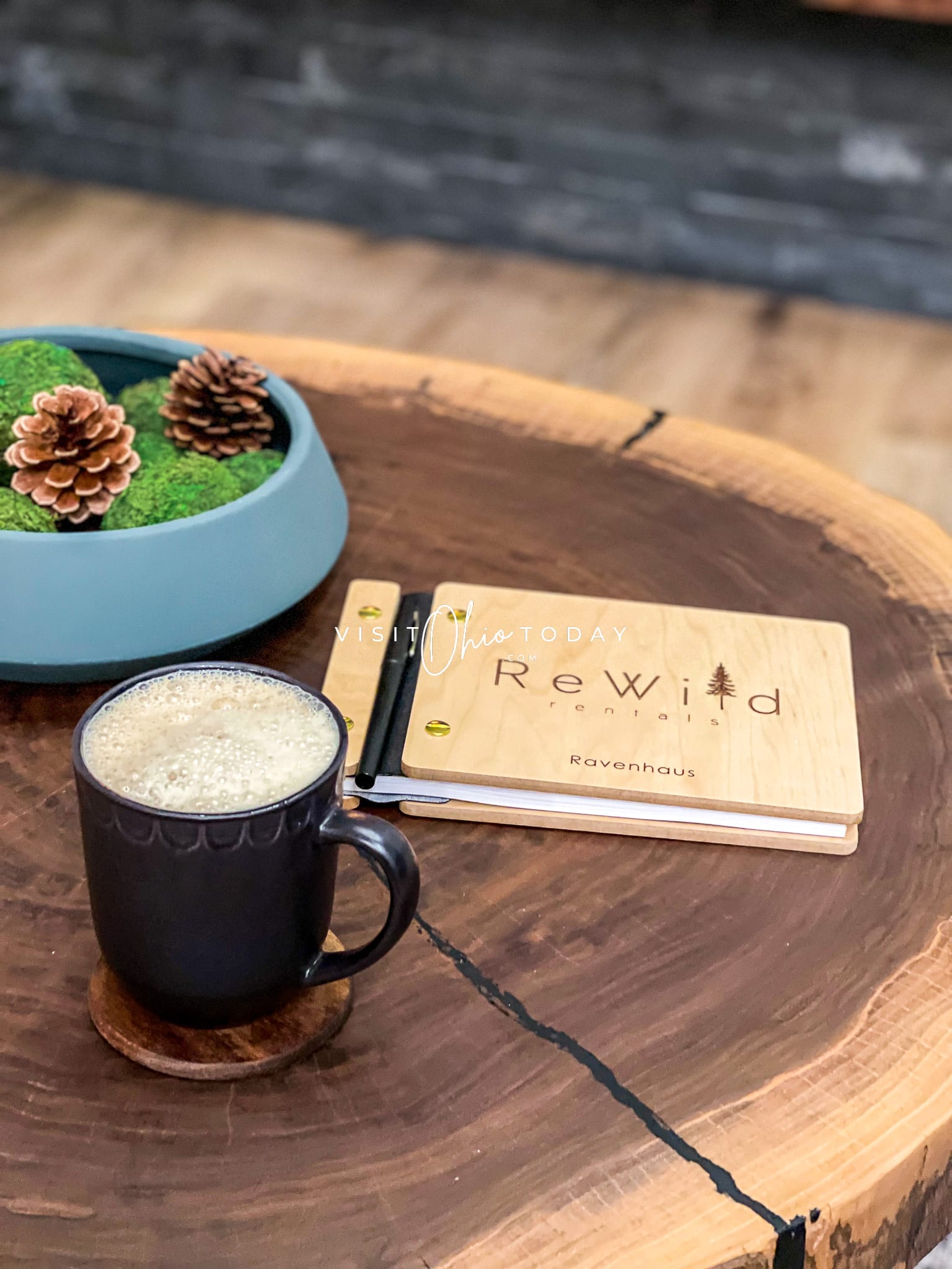 wooden table with black coffee mug filled with foamy liquid, wooden book that says rewild on it and a blue ceramic bowl filled with pinecones Photo credit: Cindy Gordon of VisitOhioToday.com