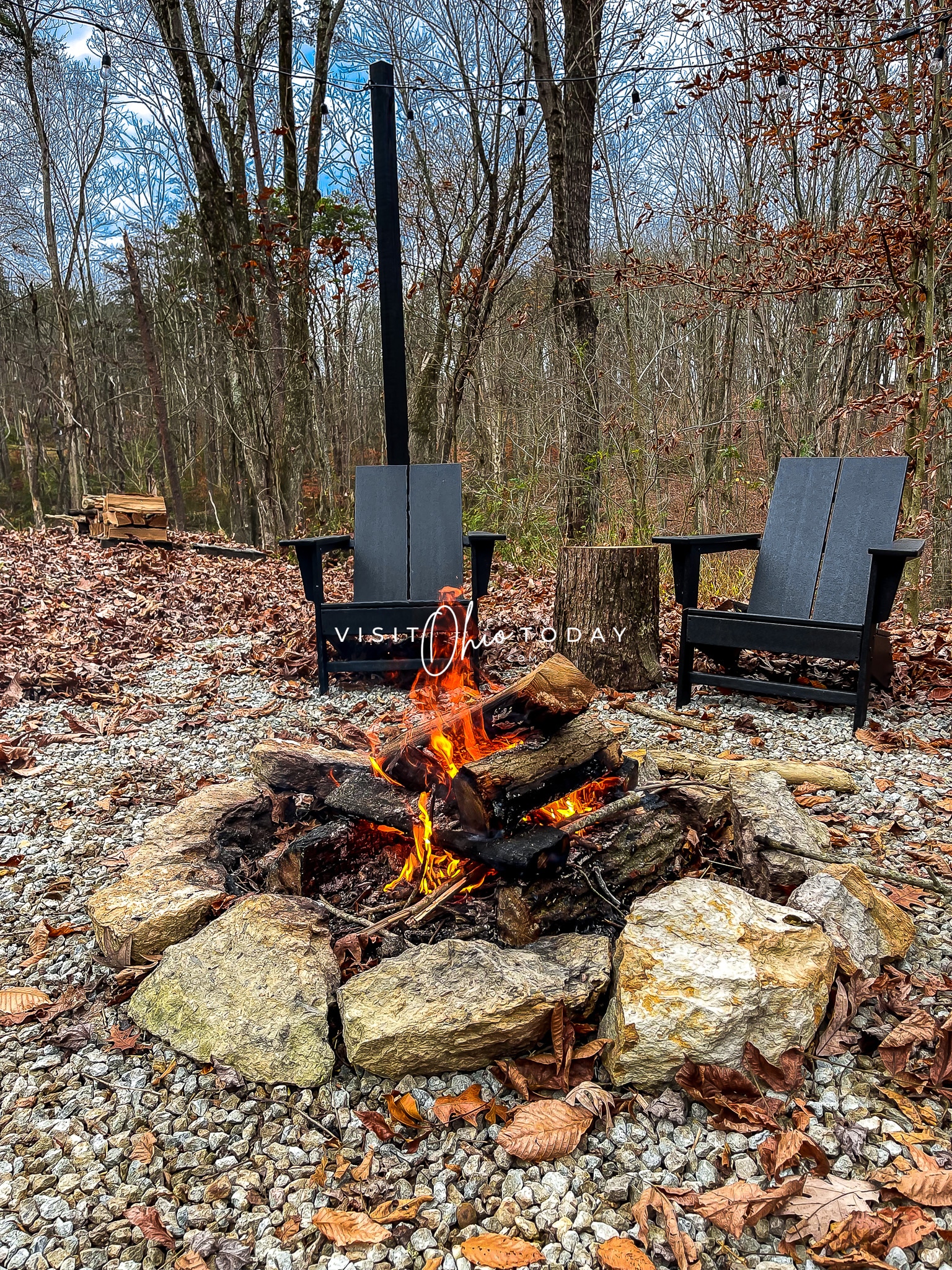 outside in a forest, fire going and two black chairs in background
