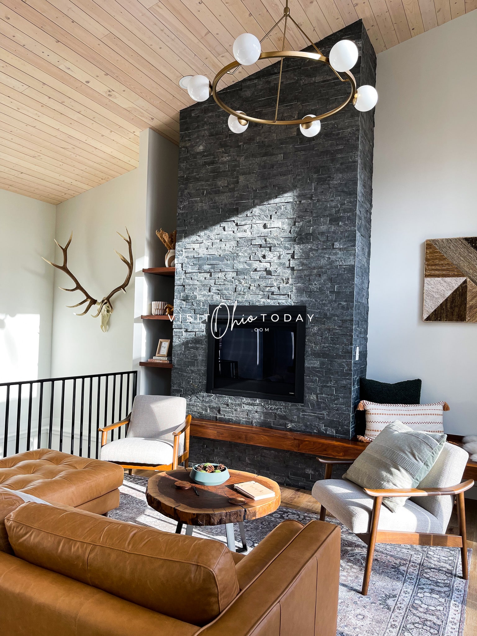 living room with brown leather cough and two white chairs with wood circle coffee table. Gas fireplace in a gray stone surround that goes up the wall, deer antlers on the left Photo credit: Cindy Gordon of VisitOhioToday.com