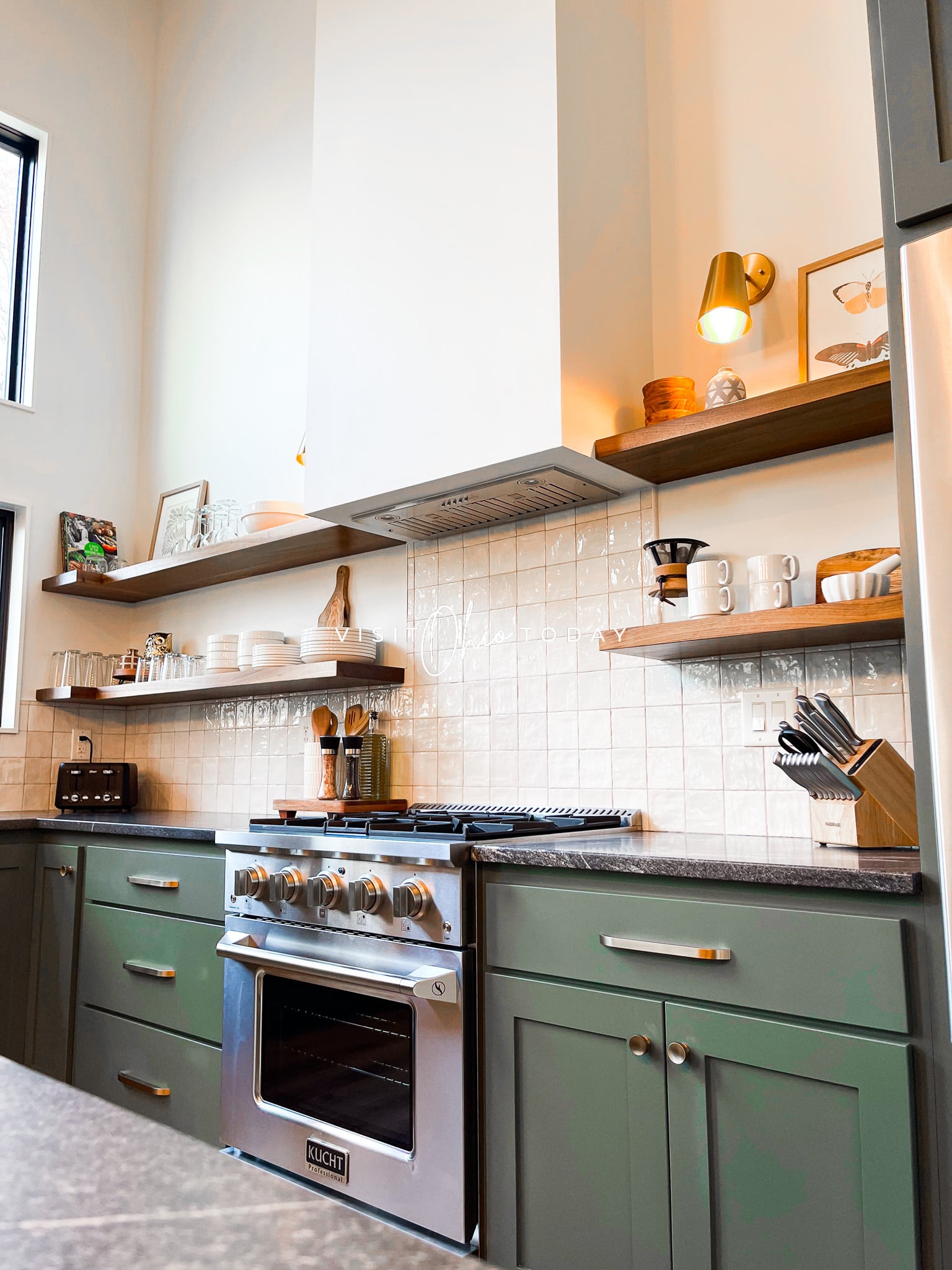 kitchen with white and tileed white wall, green cabinets, black counter tops, silver gas stove in middle and floating brown wooden shelves Photo credit: Cindy Gordon of VisitOhioToday.com