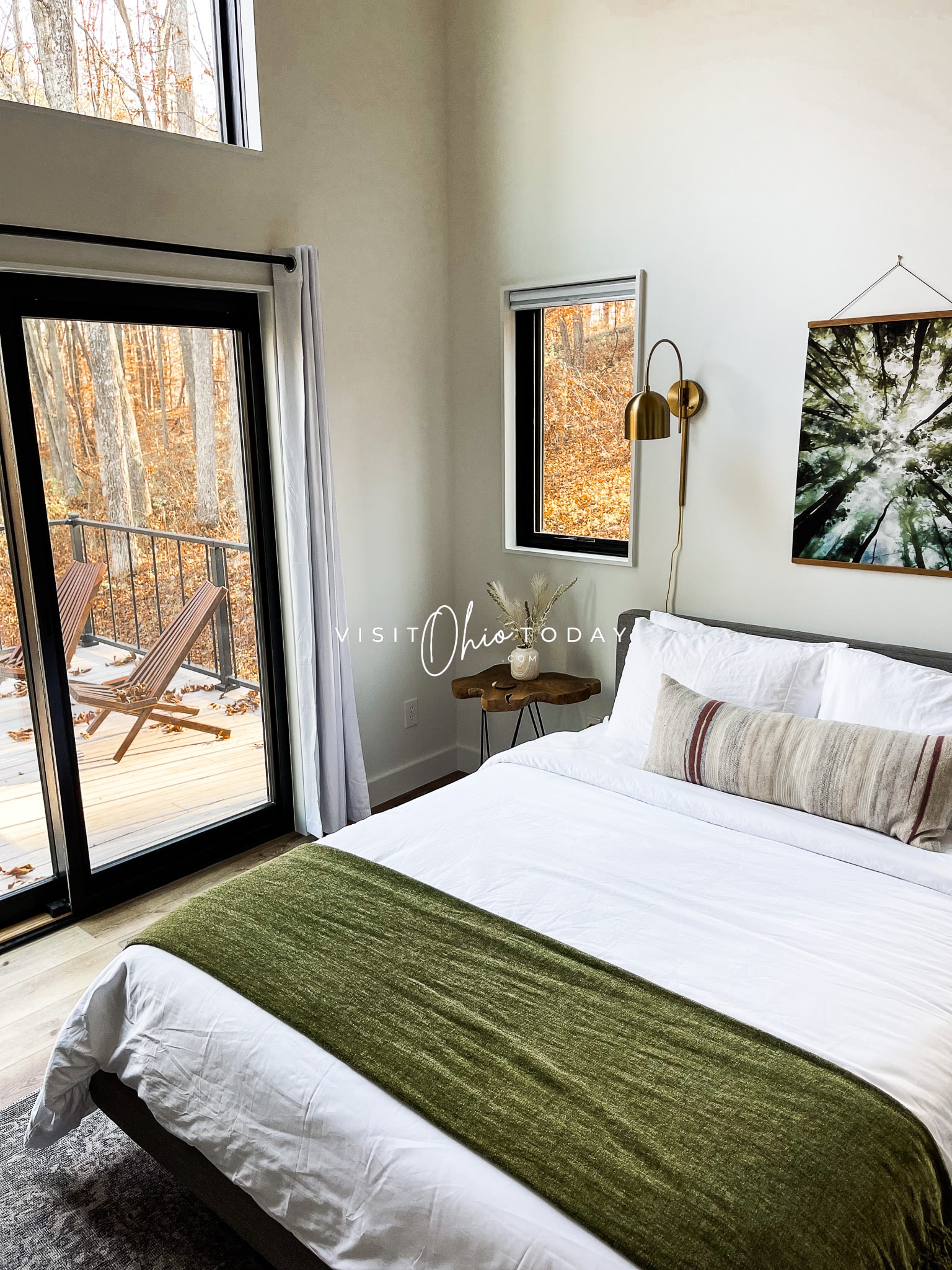 owners suite at ravenhaus, white bed with green blanket folded at end. Pillows on bed, frame on wall, windows that show forest outside Photo credit: Cindy Gordon of VisitOhioToday.com