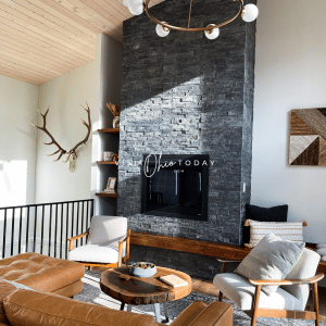 living room with brown leather cough and two white chairs with wood circle coffee table. Gas fireplace in a gray stone surround that goes up the wall, deer antlers on the left
