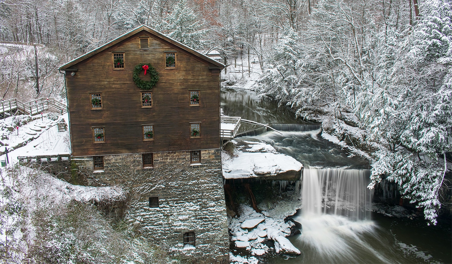 horizontal photo of Lanterman's Mill in the snow at Christmas, with a large wreath on the side of the building and smaller wreaths in each window. Image via Wikimedia Commons