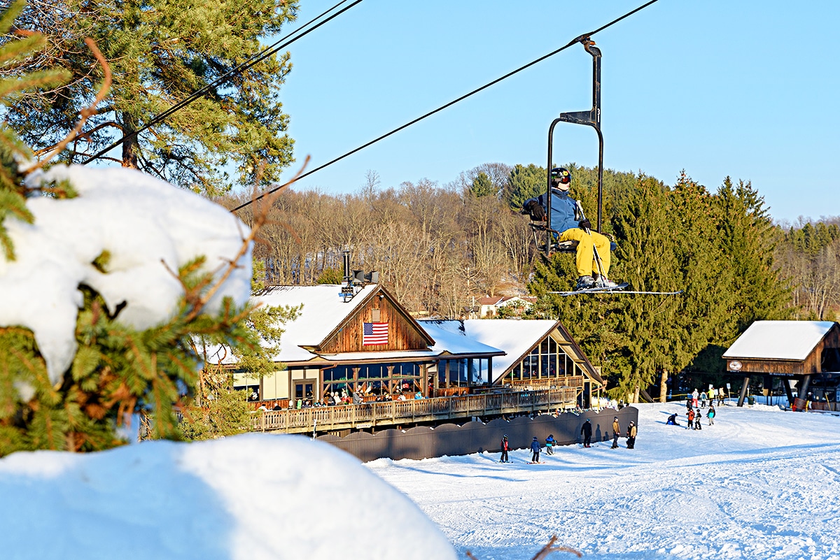 horizontal image of a skier on a ski lift with the Snow Trails building in the background