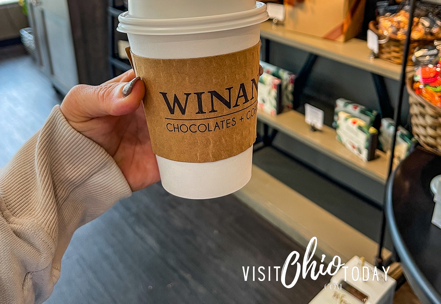horizontal photo of a hand holding a cup of Winans Coffee with a display counter in the background. Photo credit: Cindy Gordon of VisitOhioToday.com