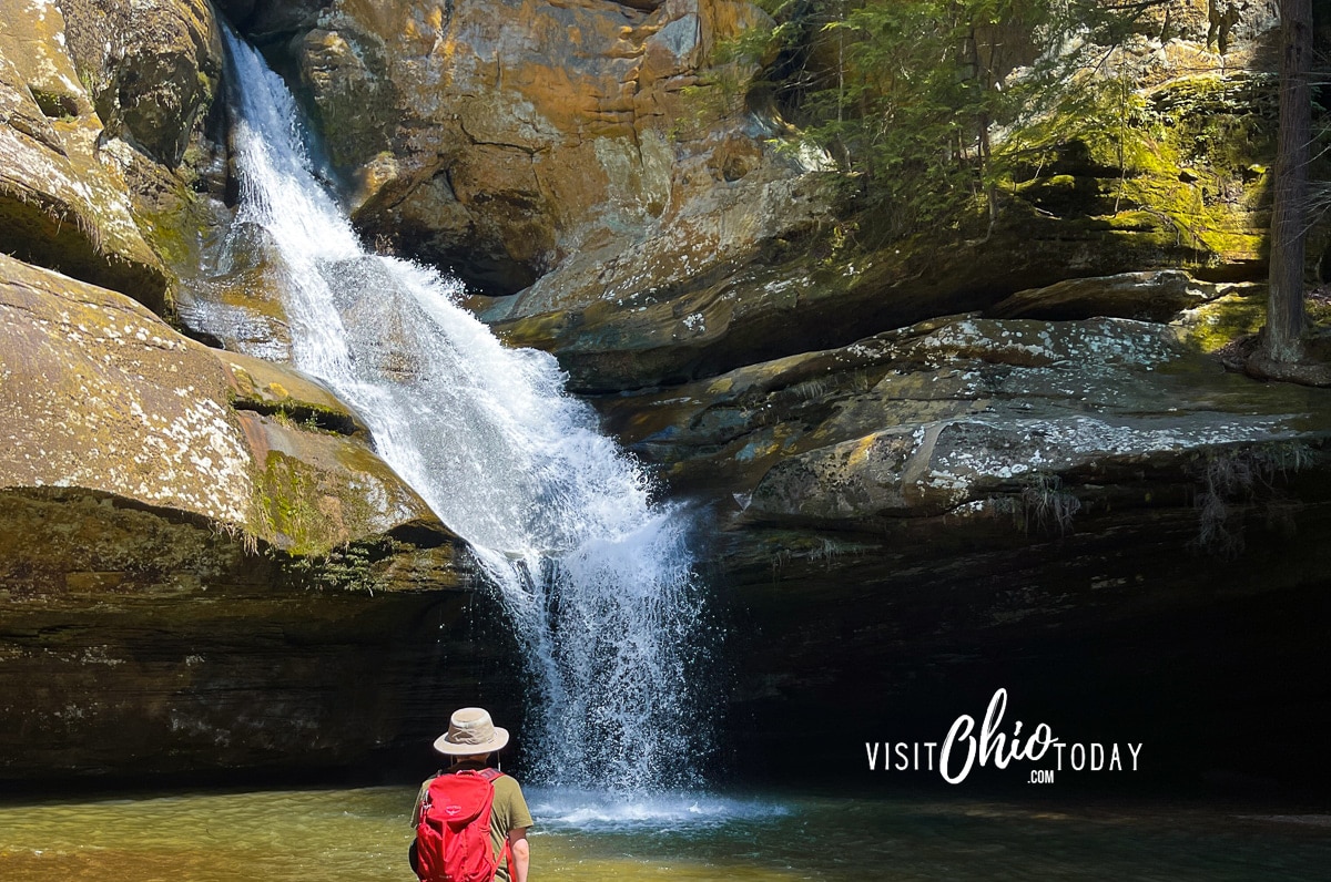 unobstructed view of cedar falls waterfall, water flowing down rocks into a clear pool, person with shorts, a red backpack and hat standing facing the waterfall Photo credit: Cindy Gordon of VisitOhioToday.com