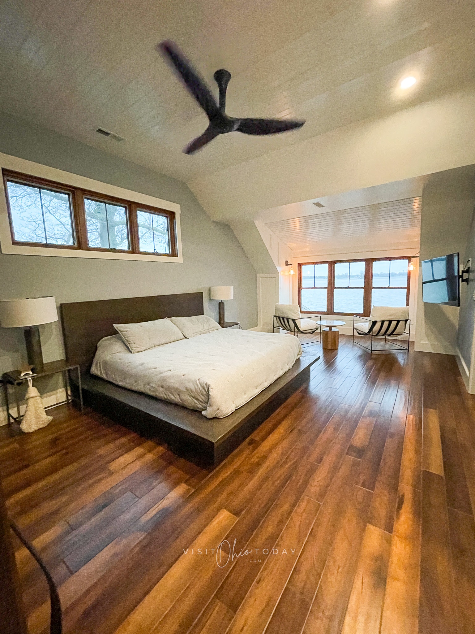 large bedroom with white ceiling and brown moving fan. large brown bed platform with white bed. Wood floors, two chairs in front of window with table inbetween. tv on right wall to side by chairs