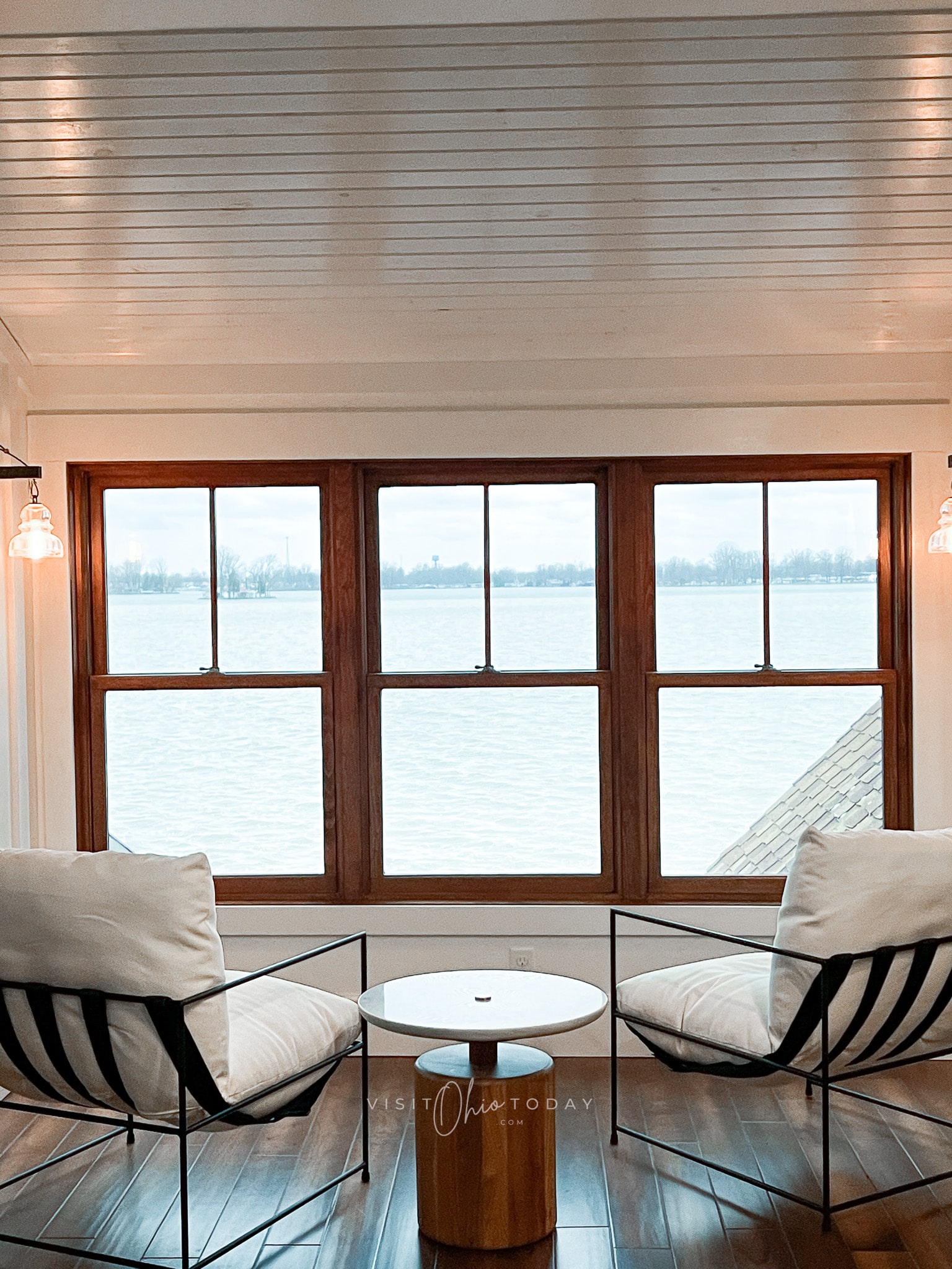 white celing and walls, three windows looking out to lake. two black chairs with white cushions in front of window
