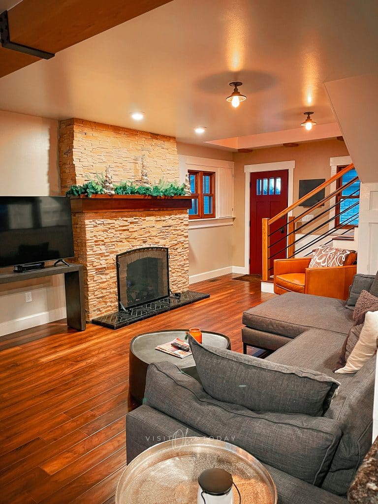 stone tiled fireplace with black fire screen. large black tv to the left of fireplace. L shaped grey couch with yellow leather chair and a tear drop shaped coffee table in front of couch
