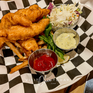 white plate with black and white checked paper on it. Breaded fish strips, sauces and coleslaw