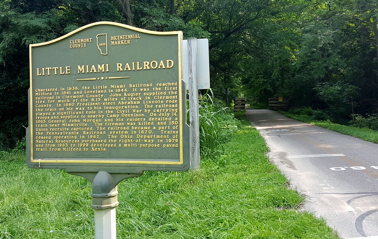 horizontal photo of part of the little miami scenic trail with a paved part of the trail, a grass verge and a bicentennial marker sign for the little miami railroad
