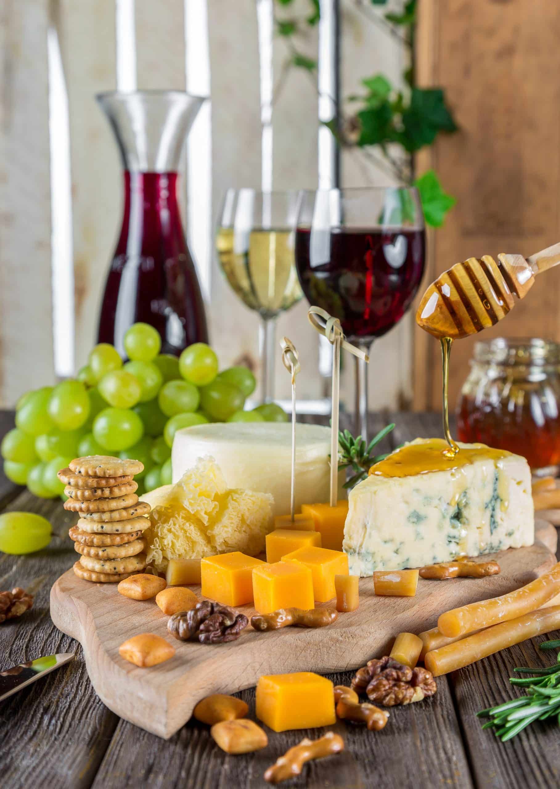 A selection of cheeses, crackers, grapes and honey on a cutting board. Glasses of red and white wine are in the background