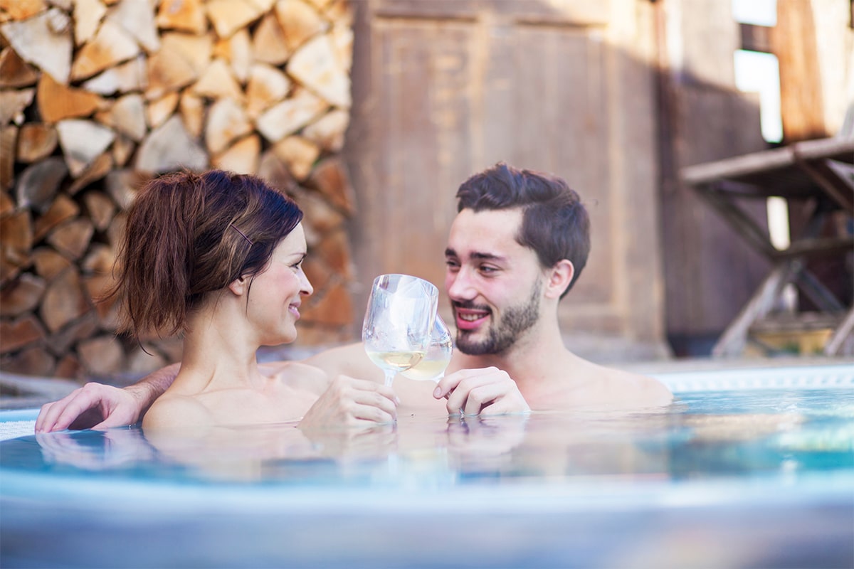 horizontal photo of a man and woman in a hot tub with glasses of champagne. A log pile is in the background. Image via Canva pro license