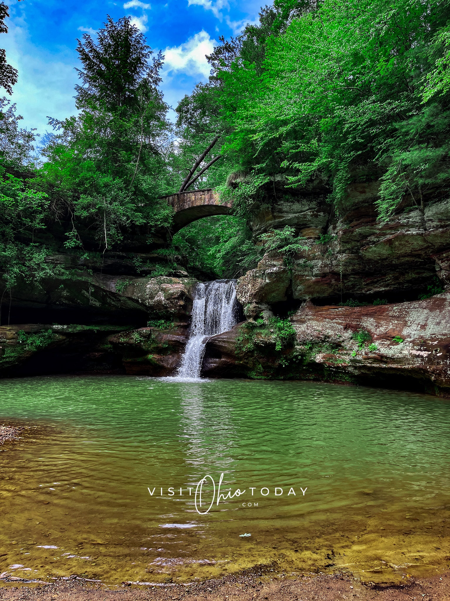 vertical photo showing the waterfall at old man's cave. Water fall flows under a stone bridge into a large pond Pond is surrounded by rocky walls. 