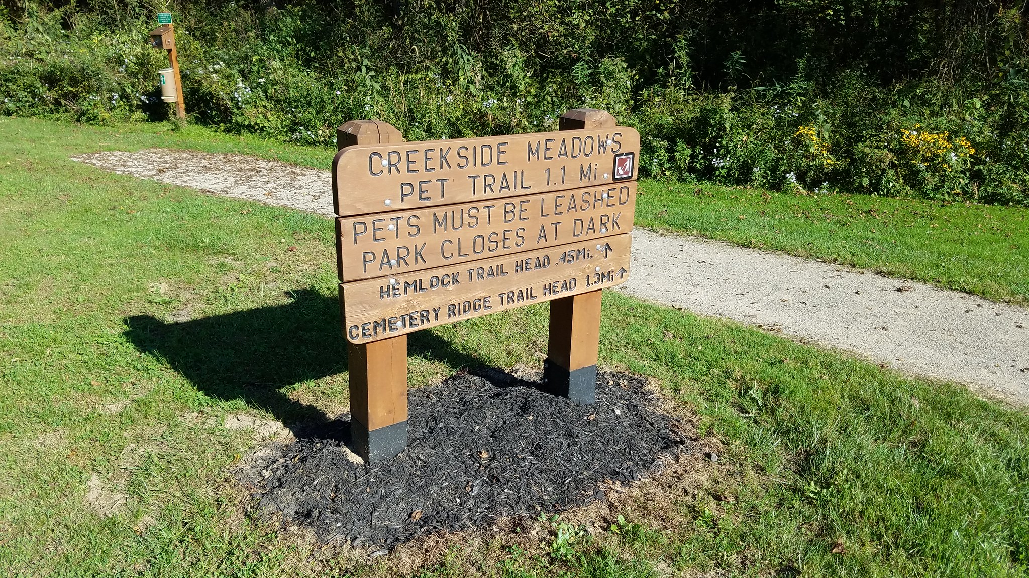 horizontal photo showing a trails sign in clear creek metro park, listing creekside meadows pet trail, hemlock trail and cemetery ridge trail. A wooden sign on a grassy area with plants and trees in the background