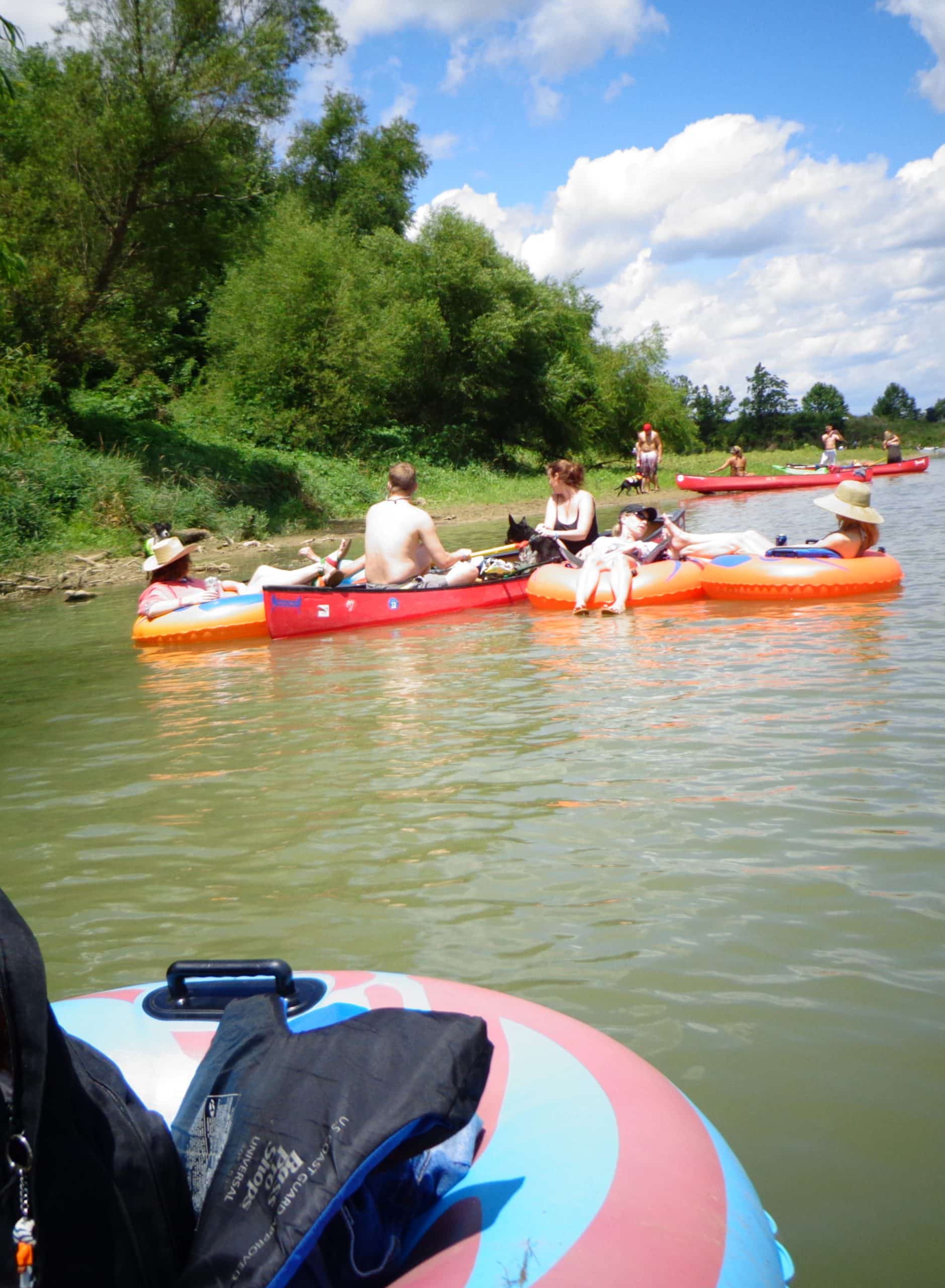 vertical photo showing a group of people in river tubes, dinghies and canoes by the bank of a river that has trees and foliage. Part of a river tube is in the foreground with a backpack on it