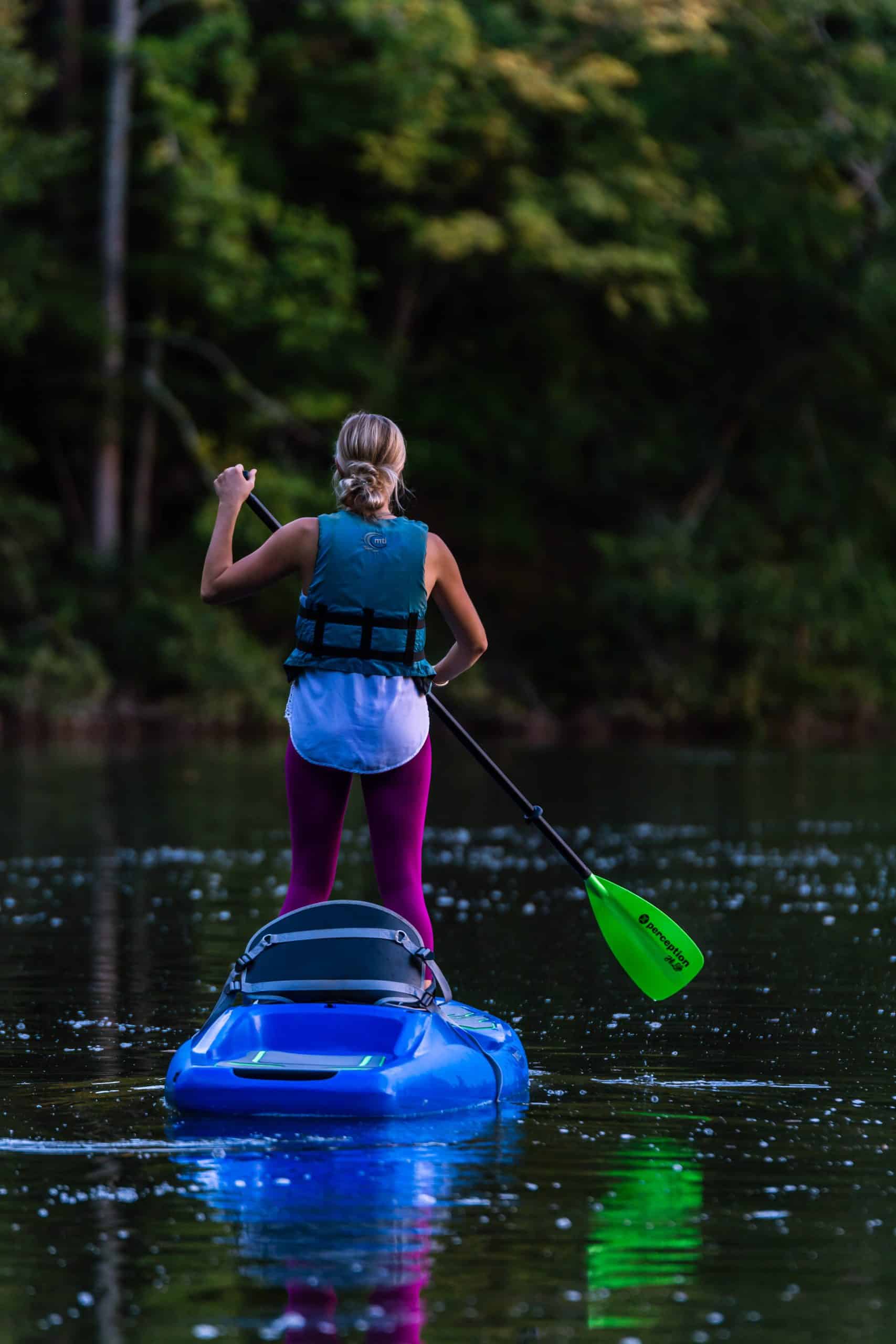 vertical photo of a woman standing in a blue  kayak with trees in the background