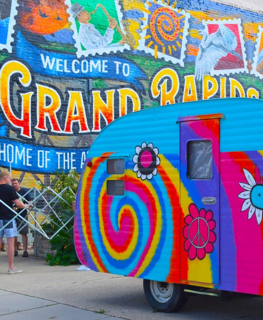 Vertical photo showing a very colorful caravan in front of a colorful hoarding with Welcome to Grand Rapids on it (fall festivals in ohio)