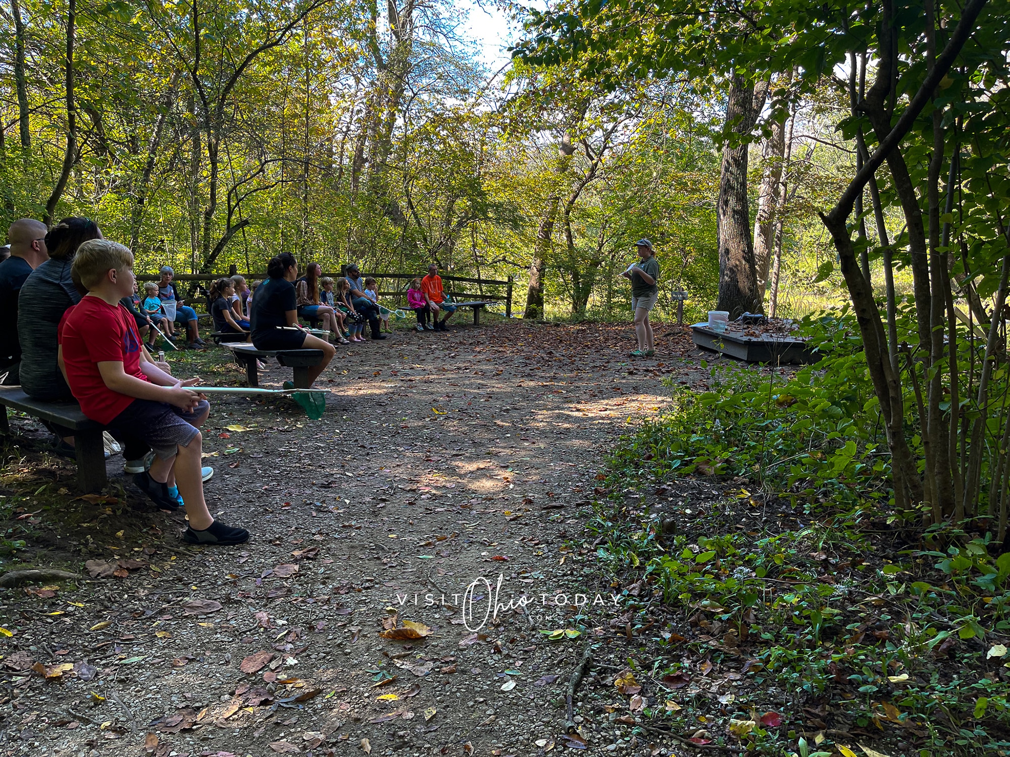 horizontal photo showing an educational session at battelle darby creek metro park with people sitting at tables in a picnic area and someone doing a talk